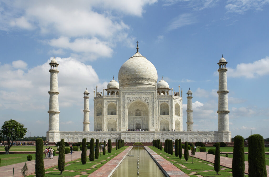 The iconic Taj Mahal with its shimmering white marble dome, standing symmetrically with minarets at each corner, surrounded by lush green gardens.