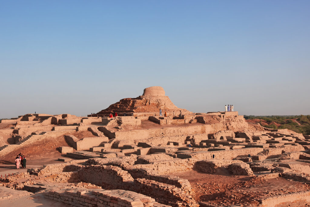 A panoramic view of Mohenjo-Daro ruins, showing wide, intersecting streets and ancient building foundations.